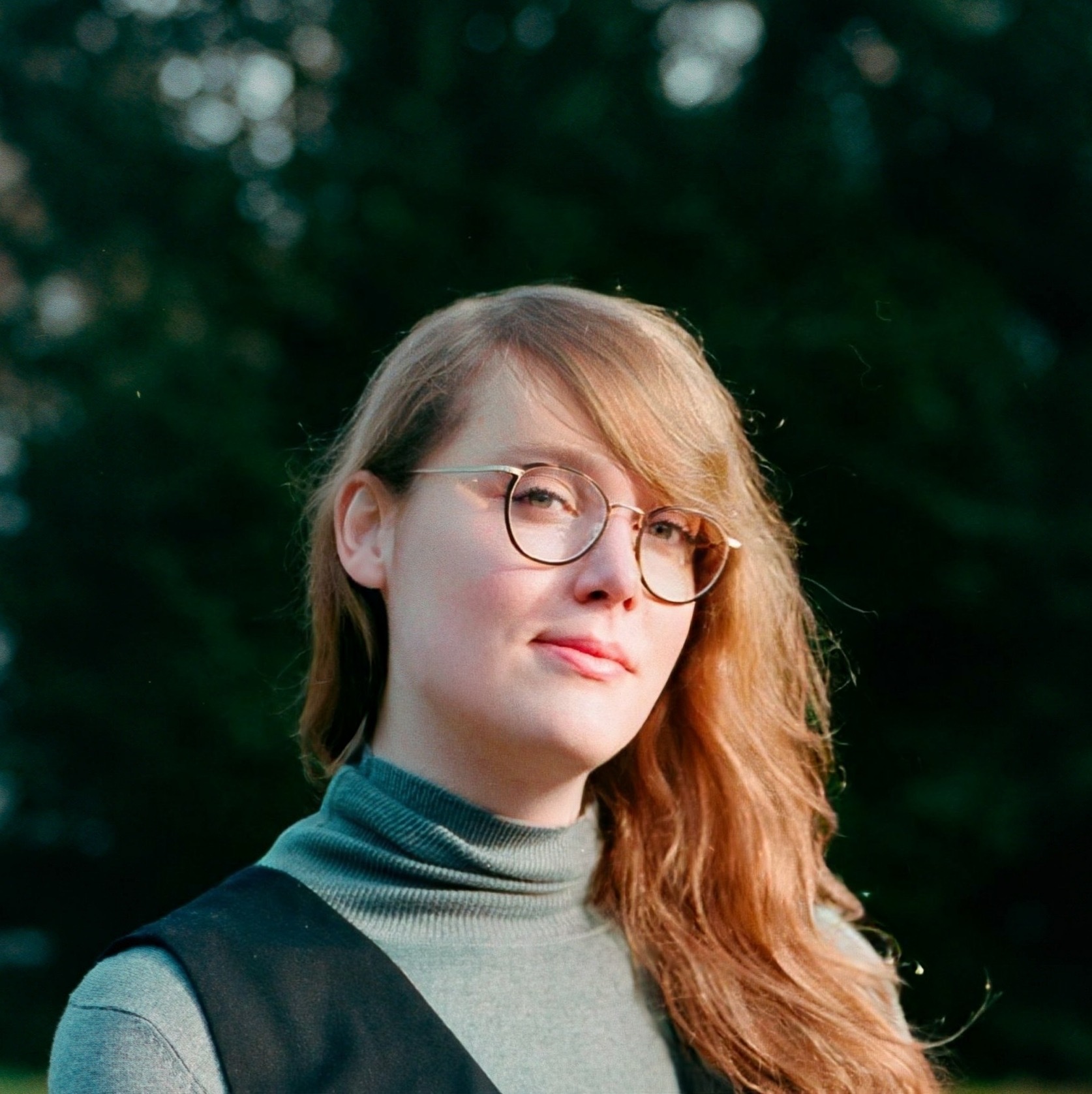 Mare met lang haar en een bril staat buiten. De persoon draagt een trui met een col en een zwart hesje. De achtergrond is onscherp met groene bomen. De persoon kijkt naar de camera.