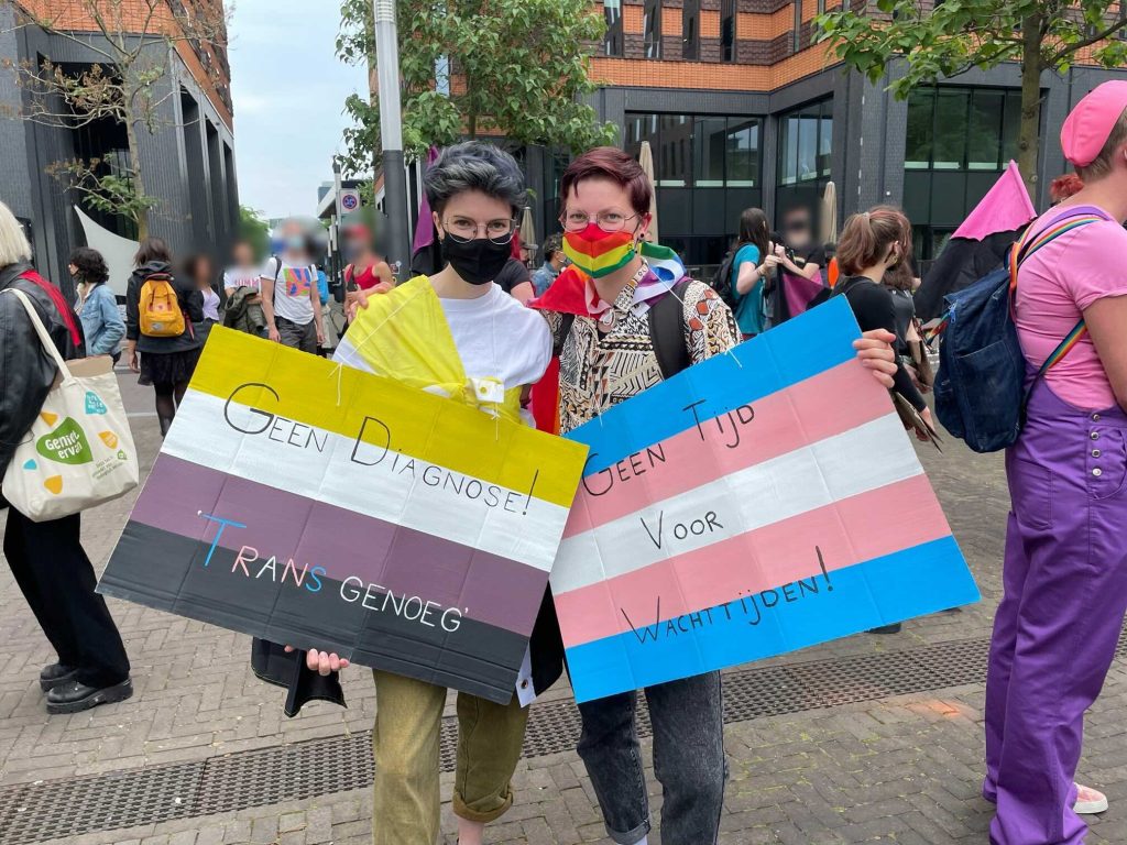 foto van 2 personen bij demonstratie met demo bordjes met de teksten: Geen Diagnose! Trans genoeg en Geen Tijd voor Wachttijden!