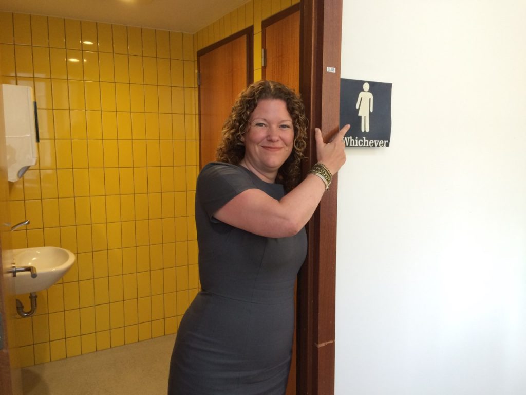 foto van wc met genderneutraal bordje en vrouw ernaast die er met glimlach naar wijst
