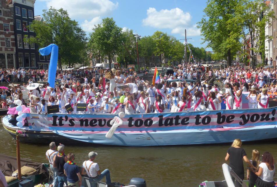 foto van de transboot tijdens de Canal Parade Amsterdam met mensen met witte kleren en roze en blauwe sherps en de tekst It's never too late to be you!