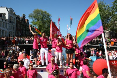 Foto van een boot op de Canal Parade Amsterdam met mensen met roze t'shirts en een regenboogvlag.