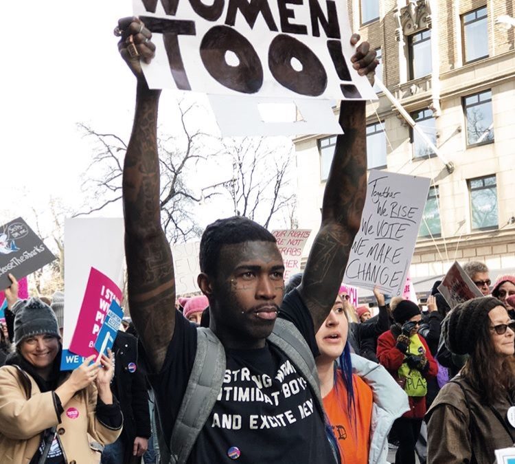 Foto van veel mensen tijdens een protest met op de voorgrond een man met een bord met de tekst "Women Too!"