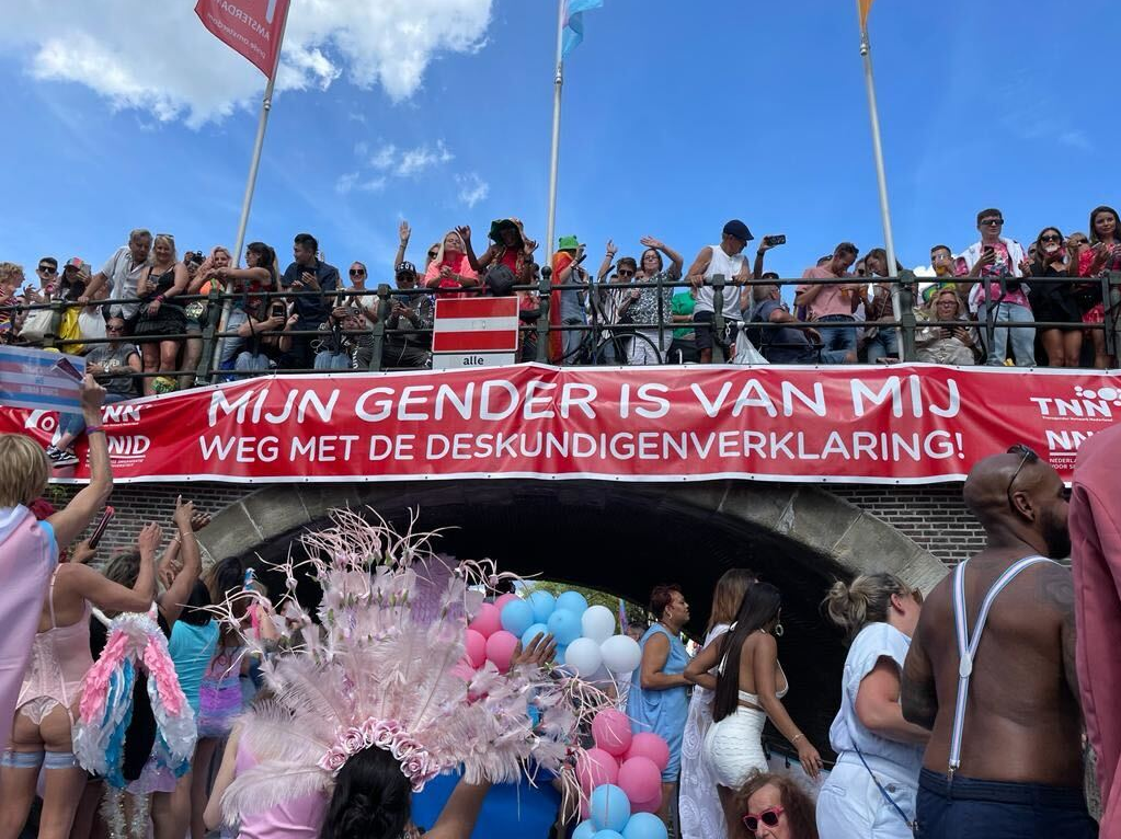 Foto van een banner met de tekst "Mijn gender is van mij, weg met de deskundigenverklaring!" vanaf een boot genomen tijdens de Pride met diverse mensen in beeld.