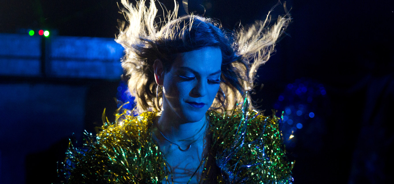 Foto van een vrouw in feestelijke kleding, waarbij haar haar omhoog wappert in de wind. De foto heeft voornamelijk blauwe accenten en er schijnt een licht achter haar voor contrast.