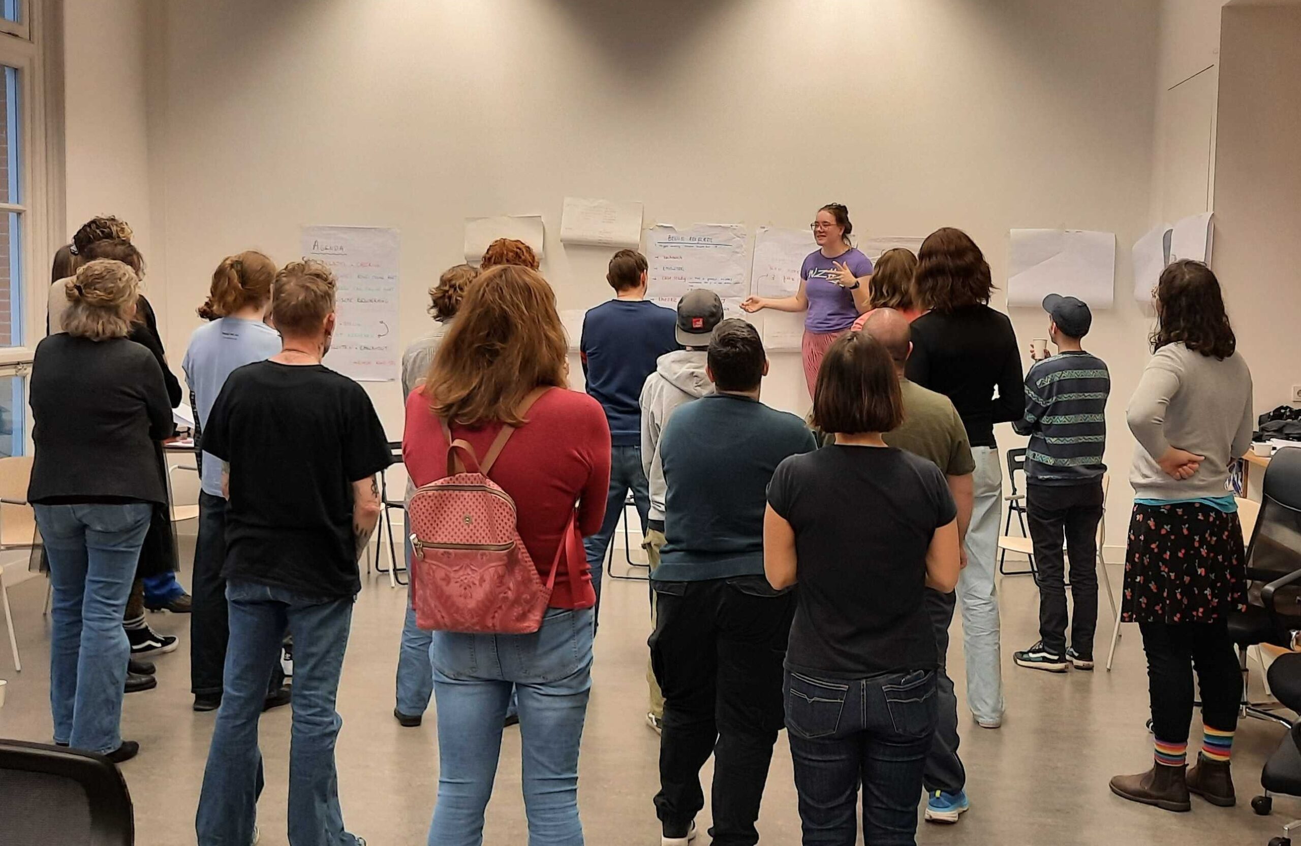 groep mensen kijkt naar de workshopleider en papieren met geschreven teksten die geplakt zijn op de muur.