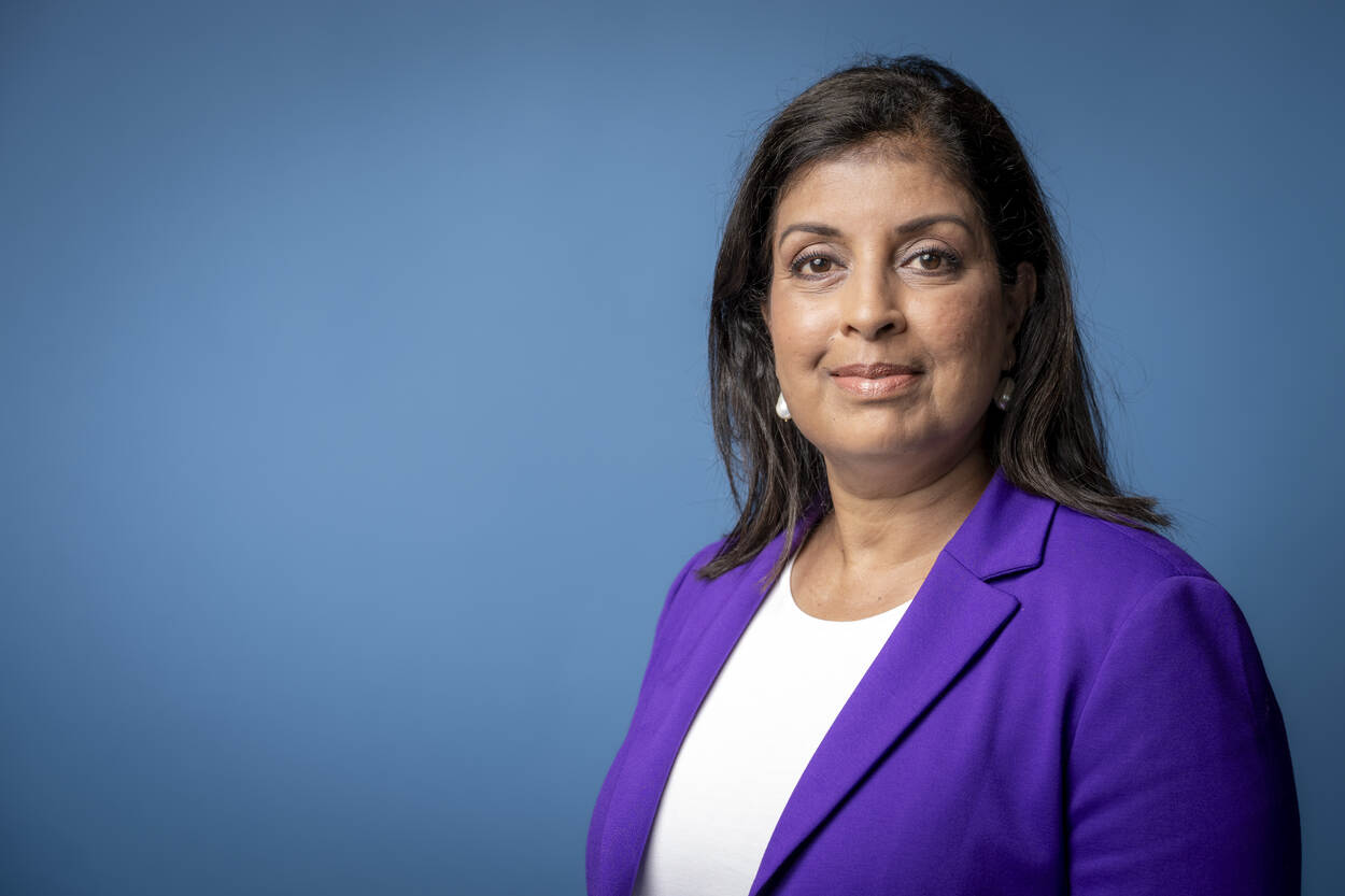 Staatssecretaris Mariëlle Paui=l, een Pakistaans-Nederlandse vrouw met een paars colbertje aan en wit shirt, staat voor een lichtblauwe achtergrond.