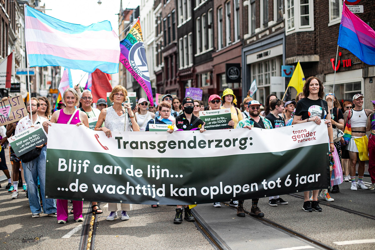 Foto van de Pride Walk, waar deelnemers demonstreren voor betere transgenderzorg. Een groep mensen loopt mee met een spandoek waarop staat: “Transgenderzorg: Blijf aan de lijn… de wachttijd kan oplopen tot 6 jaar”, met het logo van Transgender Netwerk.