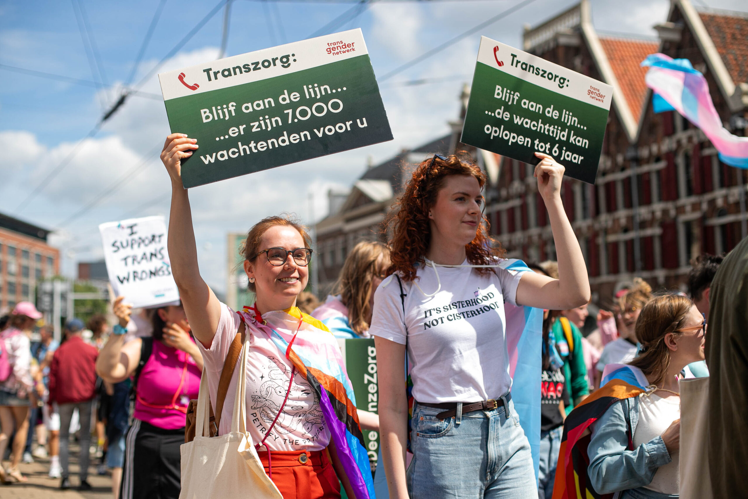 Twee deelnemers van de Trans Pride Walk 2025 tonen protestborden van Transgender Netwerk. De persoon links van het duo draagt een bord met de tekst “Blijf aan de lijn… er zijn 7.000 wachtenden voor u.” De persoon rechts van het duo draagt een bord met de tekst “Transgenderzorg: Blijf aan de lijn… de wachttijd kan oplopen tot 6 jaar” en een t-shirt met de tekst “sisterhood not cisterhood” Op achtergrond zijn andere demonstranten te zien waarvan één een bord draagt met de tekst “I support trans wrongs”