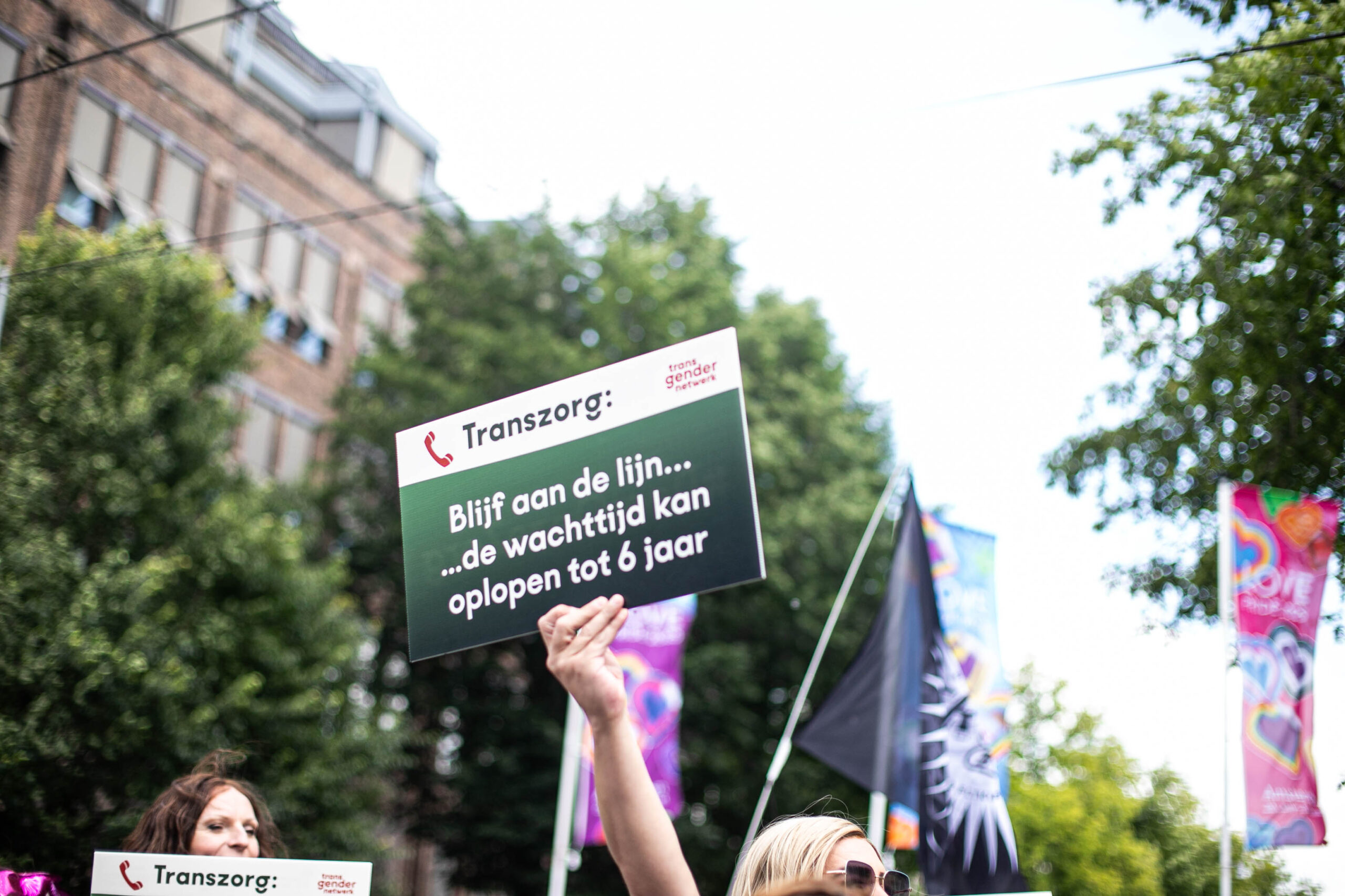 Een protestbord bij Trans Pride Walk 2025 met de tekst “Transgenderzorg: Blijf aan de lijn… de wachttijd kan oplopen tot 6 jaar” en het logo van Transgender Netwerk.