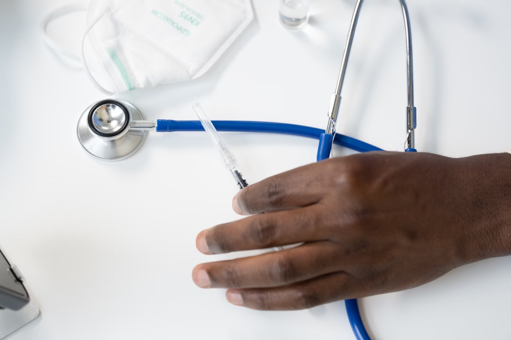 Close-up van een hand op een bureau met een stethoscoop, injectiespuit en mondmasker, symbool voor medische zorg en behandeling