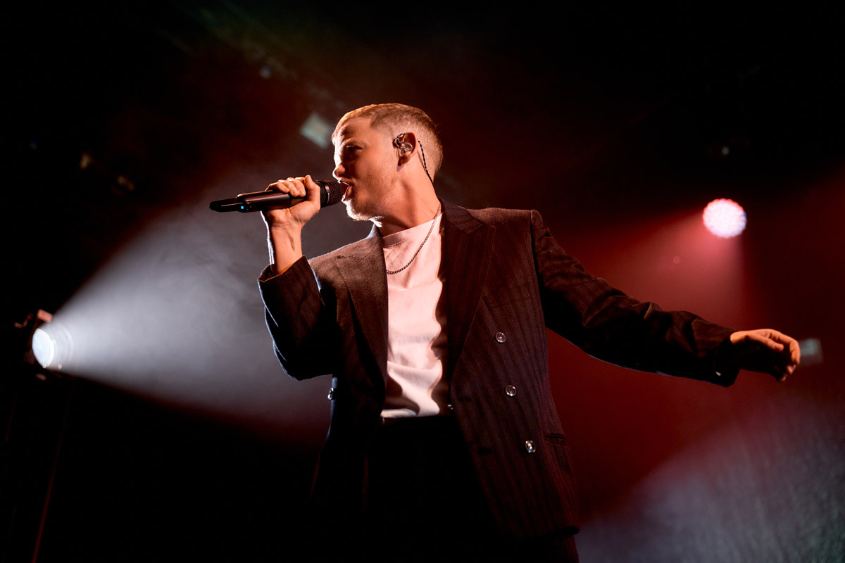 Kae Tempest in een donker pak en wit shirt zingt in een microfoon op een podium. Er is fel licht op de achtergrond. De persoon draagt een oorstukje en een ketting, en houdt de andere arm wijd uit. De sfeer is energiek en professioneel.