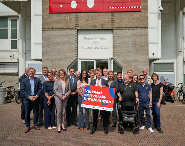 Een groep mensen staat voor het gebouw van de Eerste Kamer der Staten-Generaal. Ze houden een groot bord vast met de tekst: Verbied conversiehandelingen! De groep bestaat uit een mix van personen, waaronder mensen in formele kleding en een persoon met een rolstoel.