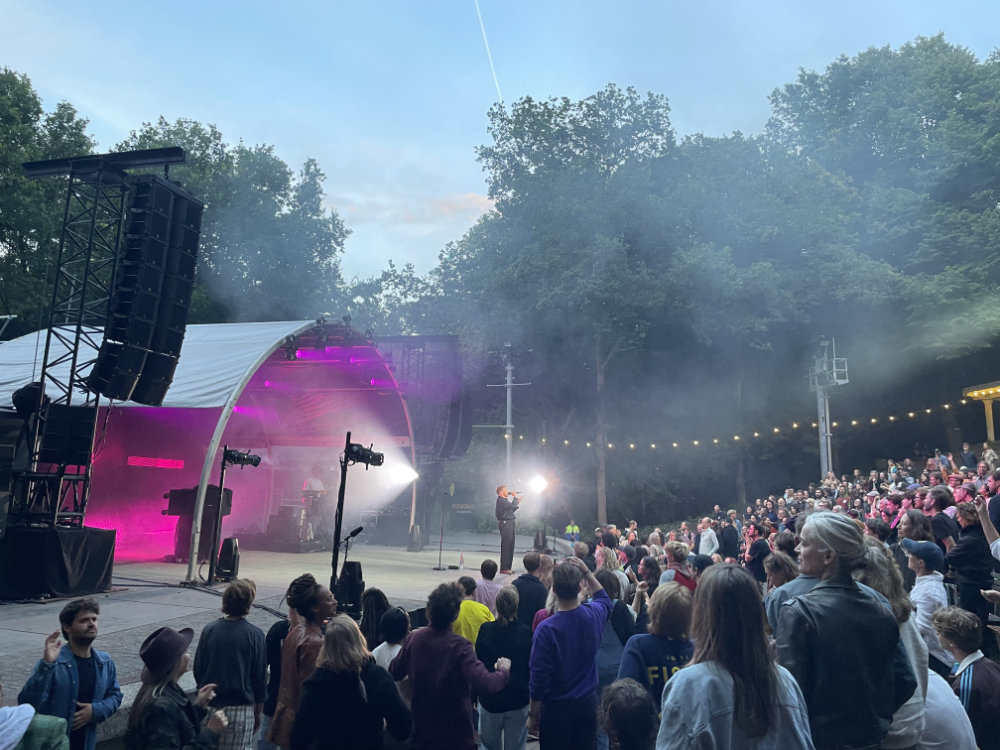 Openluchtconcert bij schemer: de artiest Kae Tempest staat op een verlicht podium met roze en witte lichtbundels en rook. Grote luidsprekers links. Voor het podium staat een menigte die naar het optreden kijkt. Achter het podium bomen en een rij lichtjes.