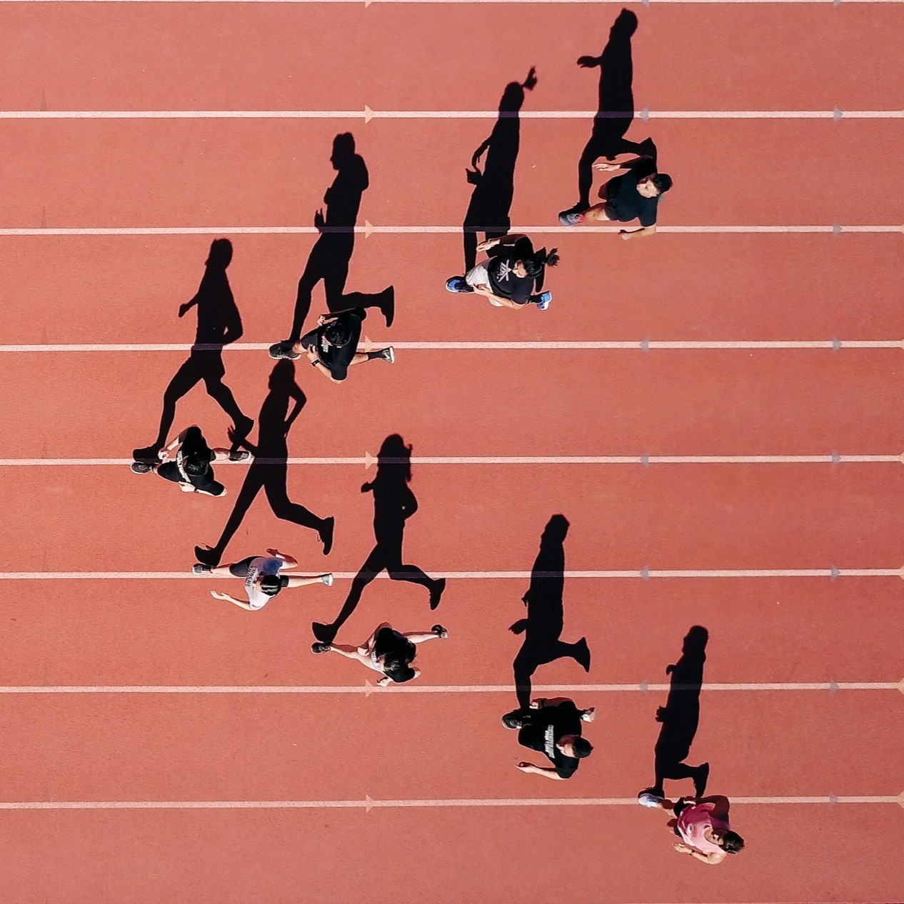 Een groep mensen rent op een rode atletiekbaan, gezien van bovenaf. Hun schaduwen zijn duidelijk zichtbaar op de baan. De baan heeft zes witte lijnen. De mensen zijn actief, mogelijk werkenden die deelnemen aan een sportevenement.