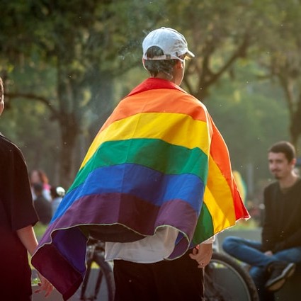 Een persoon met een witte pet heeft een regenboogvlag om de schouders en staat in een park. Op de achtergrond zitten en staan andere mensen en staan fietsen tussen bomen en groen.