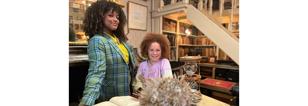 Twee personen, een volwassene en een kind, staan in een kamer met hoge boekenplanken. Op de tafel voor hen ligt een open boek en een opgeblazen stekelvis; daarnaast staan twee zandlopers. De volwassene draagt een geruit jasje.