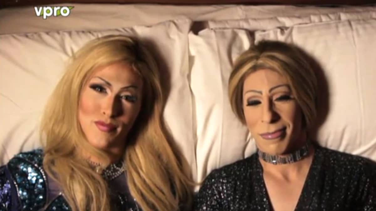 Twee personen liggen naast elkaar op een wit kussen. Ze dragen zware make-up, lange blonde pruiken, glinsterende kleding en chokerkettingen. Links kijkt iemand naar de camera. VPRO-logo linksboven.