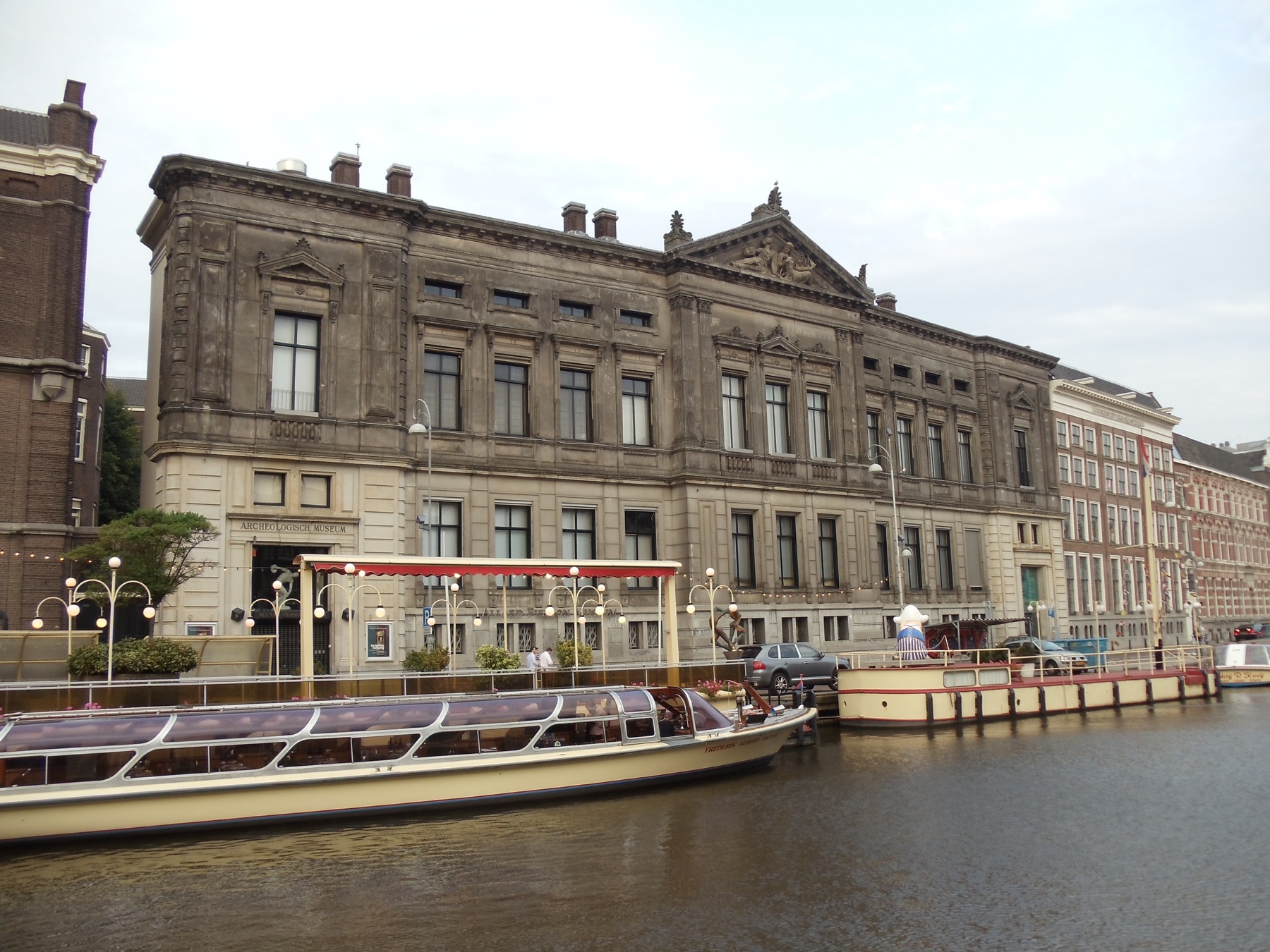Groot oud stenen gebouw met klassieke gevel en beeldengroep in het fronton. Op de gevel staat Archeologisch Museum. Voor het gebouw loopt een gracht met een glazen rondvaartboot, een kade met lantaarns en enkele mensen bij de ingang.