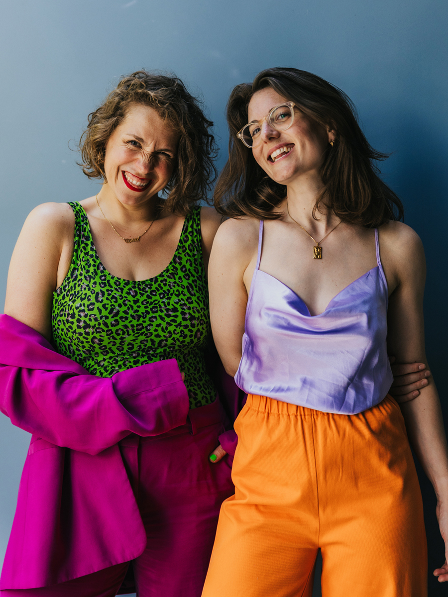 Twee personen staan dicht tegen een blauwe achtergrond, met armen om elkaar en lachend. Links draagt iemand een groene luipaardtop en fuchsia jasje en broek. Rechts draagt iemand een lila satijnen top, oranje broek en een bril.