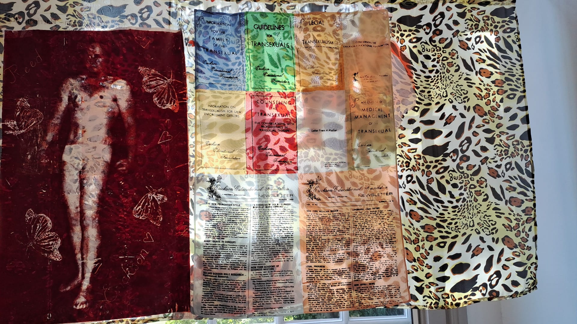Drie stoffenpanelen: links donkerrood met een witte afbeelding van een naakt persoon en vlinders; in het midden een doorzichtig paneel met gekleurde blokken en leesbare teksten over trans personen; rechts een beige stof met luipaardprint.