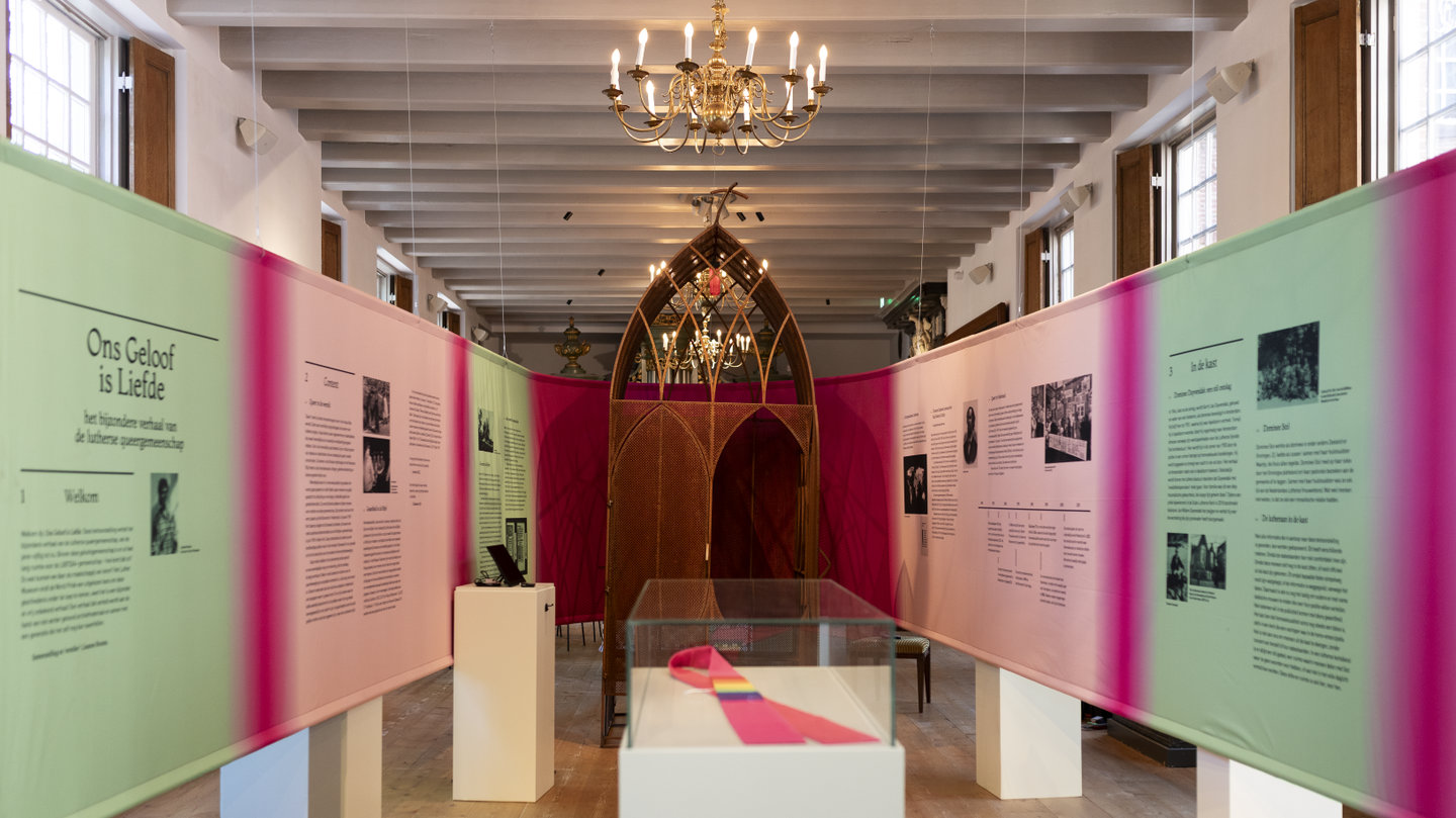 Zaal met tentoonstelling: lange roze en mintkleurige panelen met tekst langs beide zijden, een glazen vitrine met een roze sjerp op een voetstuk, en in het midden een houten boogvormig meubel onder een brandende kroonluchter.