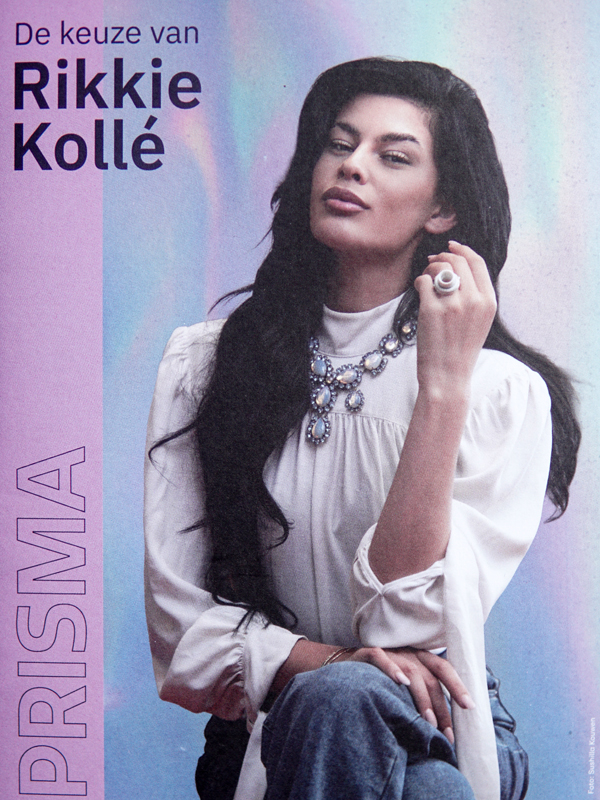 Rikkie Kollé zit in een witte blouse en blauwe spijkerbroek, met een opvallende ketting en ring en haar hand bij haar gezicht. Op de afbeelding staat ‘De keuze van Rikkie Kollé’ en ‘PRISMA’.