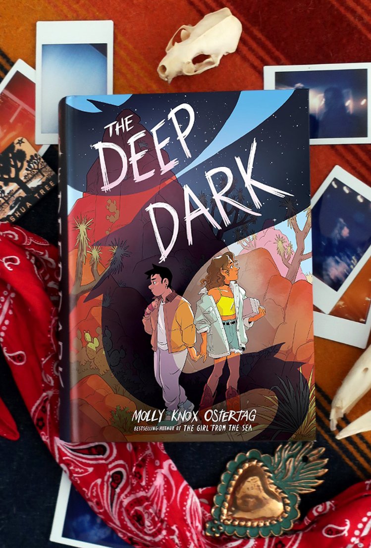 Omslag van het boek The Deep Dark met illustratie van twee jonge personen rug-aan-rug in een woestijnlandschap. Het boek ligt op een rood deken, omgeven door polaroidfoto's, een rode bandana, een dierenschedel en een groen-gouden hartvormige broche.